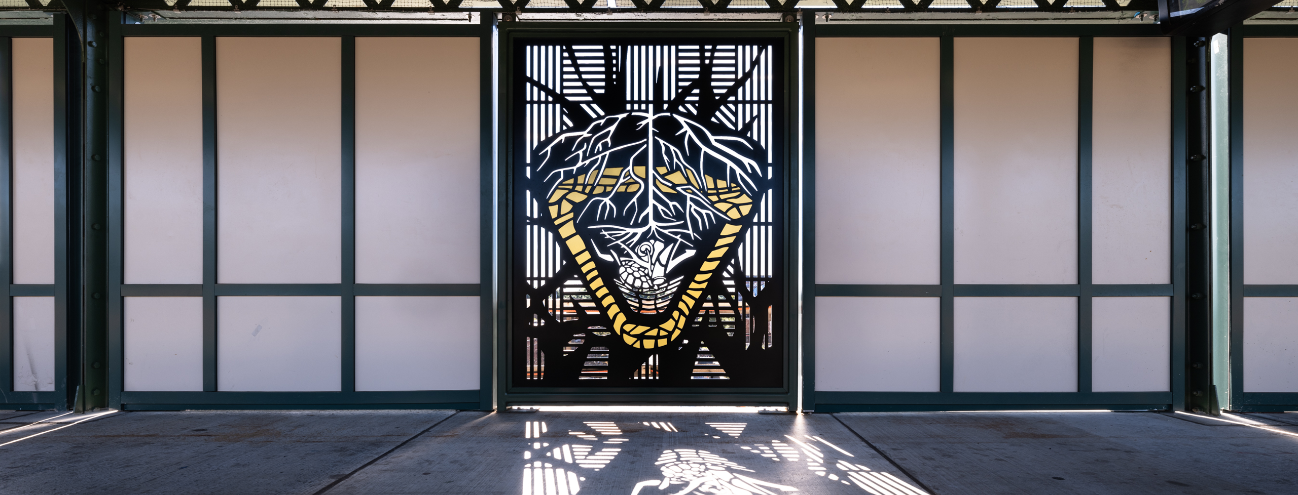 A stylized metal cut-out panel is installed in the wall of an above-ground train station platform, framed by frosted glass and dark green metal supports. The design features an earth pit with tree roots, accented by bright gold detailing. Sunlight filters through the cut-outs, casting intricate shadows onto the platform’s concrete floor below.