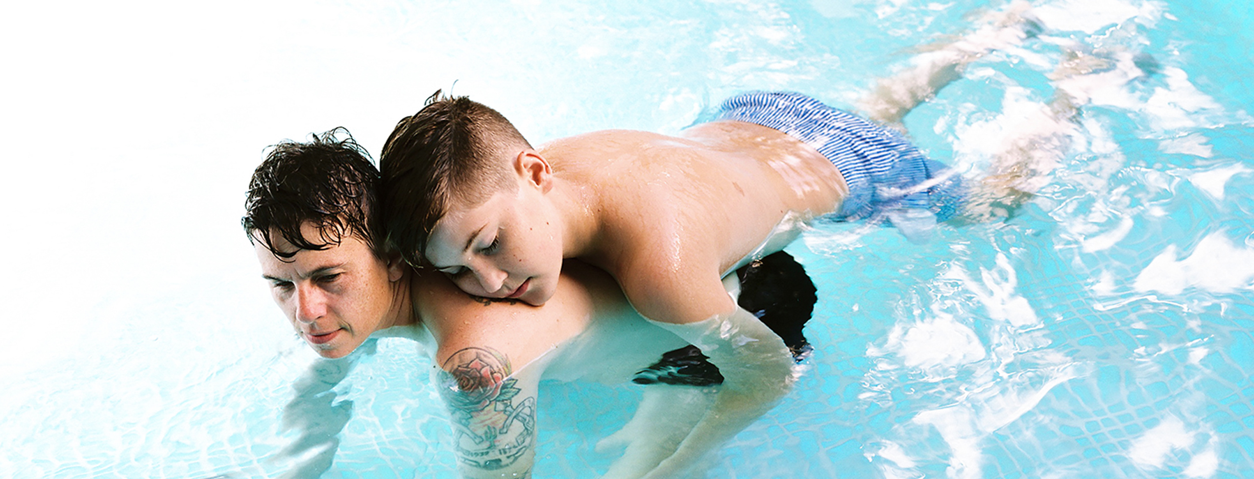A father and son are swimming in a clear pool. The son clings to his father's neck, floating on his father's back, with his eyes closed.