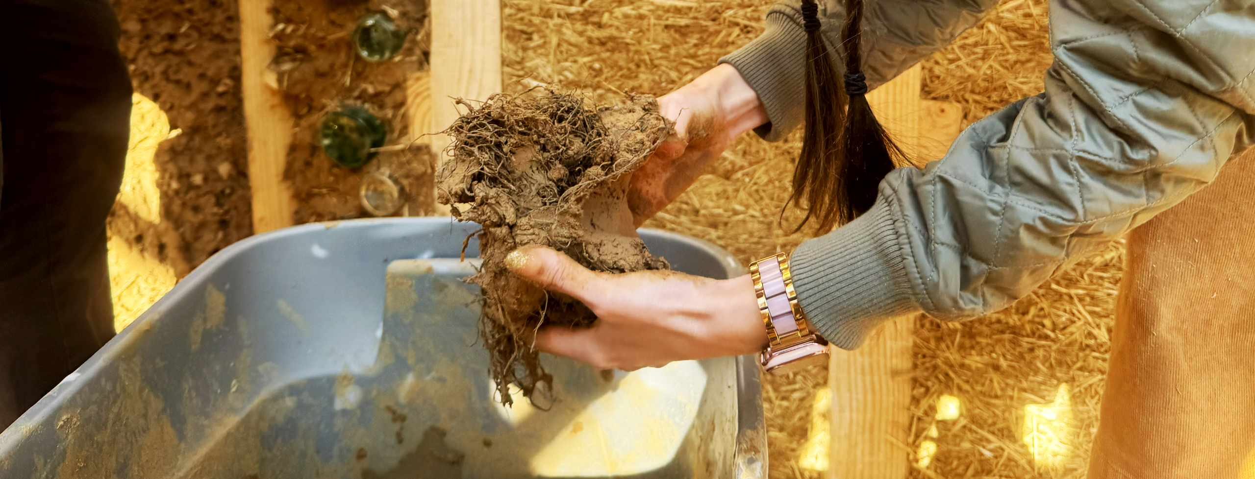 A person wearing a green quilted jacket, brown knit beanie, and long braids leans over a bin, holding a clump of clay and Spanish Moss mixture for building.