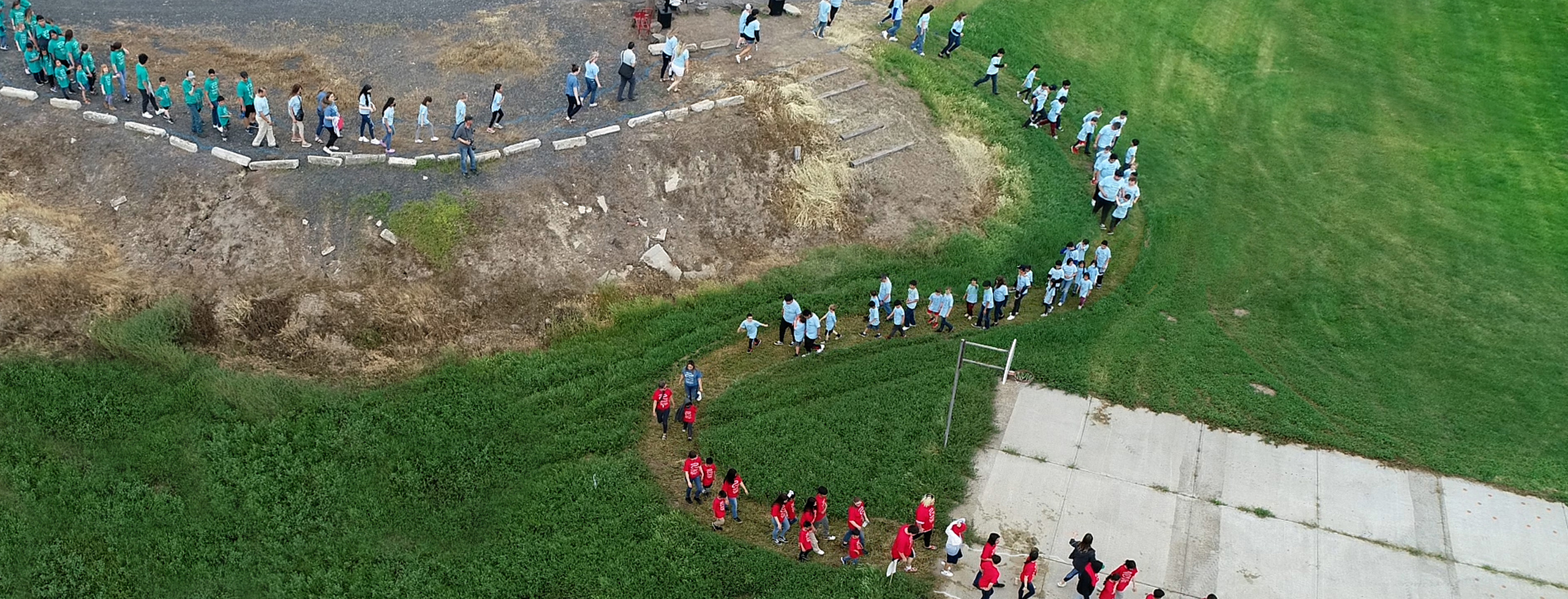 Aerial drone shot of school children of varying ages walk in pairs in a curving line in the shape of a river. The students wear matching t-shirts in turquoise, sky blue, or red that bear the project's logo.