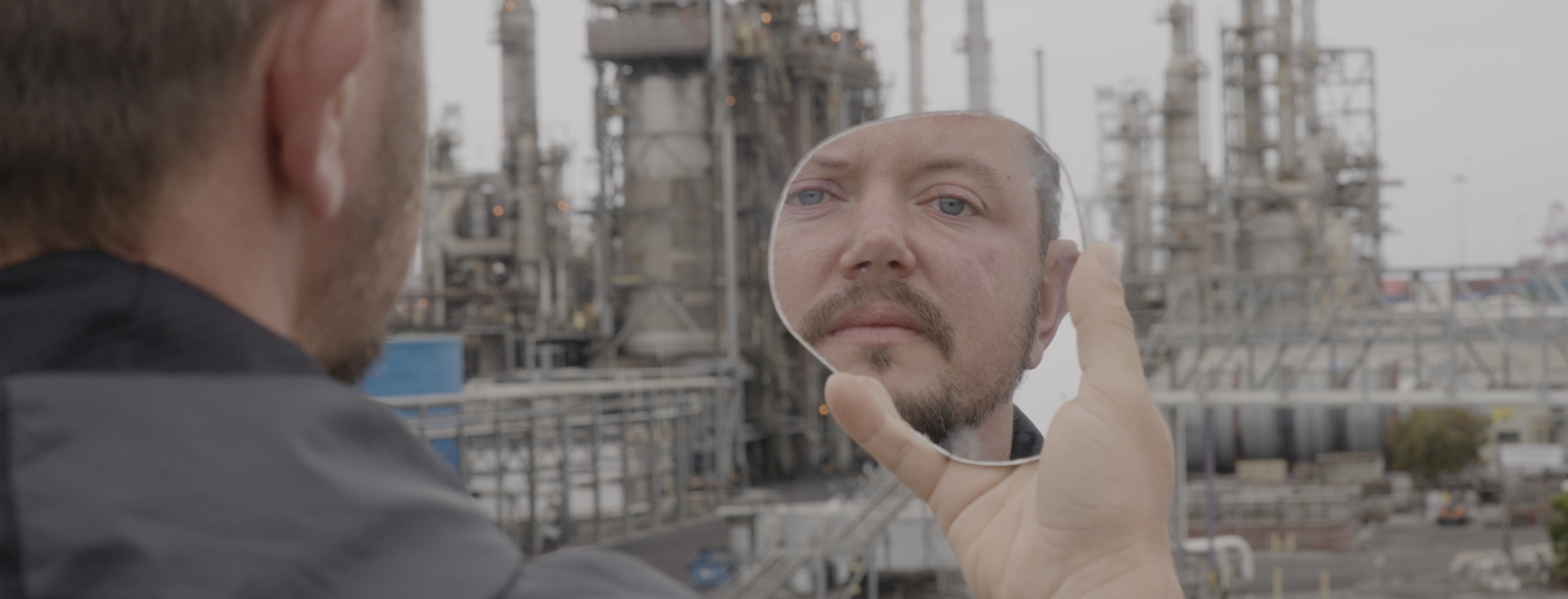 A photo of a white man from behind, standing in front of an oil refinery. His face is reflected in a small mirror he's holding up.