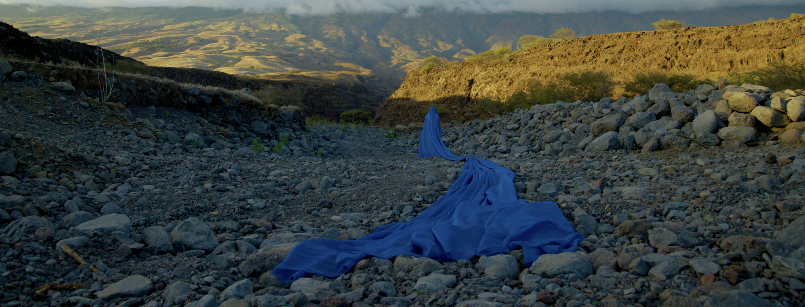 A big barren gulch of dry river rocks against a wide landscape of a mountainous region under a sky is lit by the fading light near sunset. A figure is shrouded in blue fabric, the fabric flowing along where water once flowed, as though water just emerged from a spring and has returned.