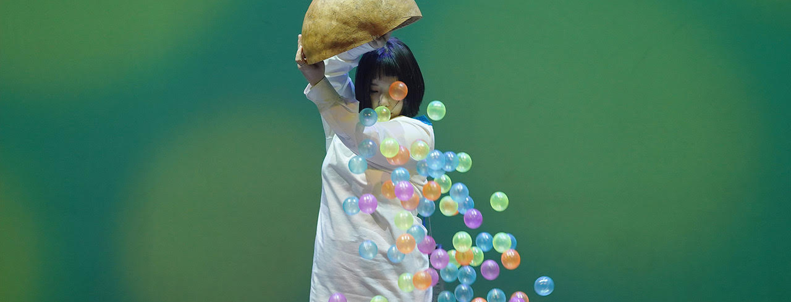 Artist Seo Jungmin pours a container of colorful plastic balls onto the floor as part of a performance, pictured against a green background