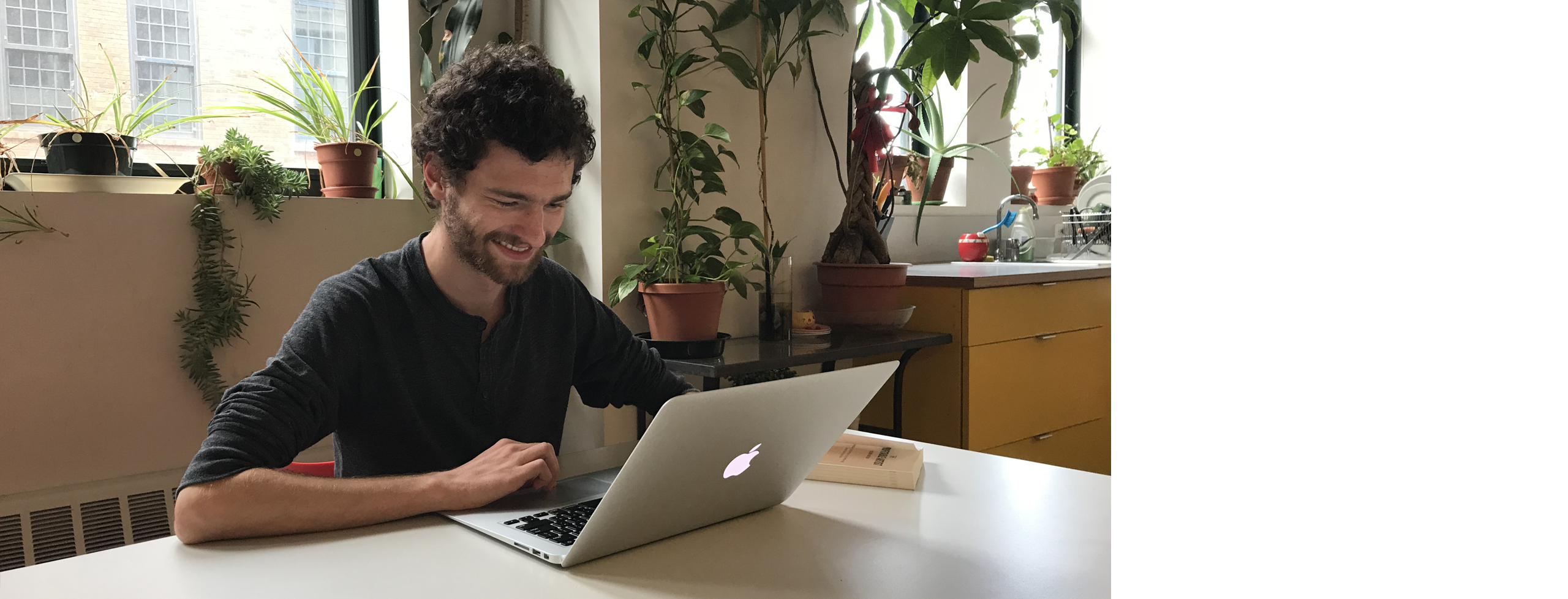 An individual sits smiling in front of a computer, light streaming in from the window behind them