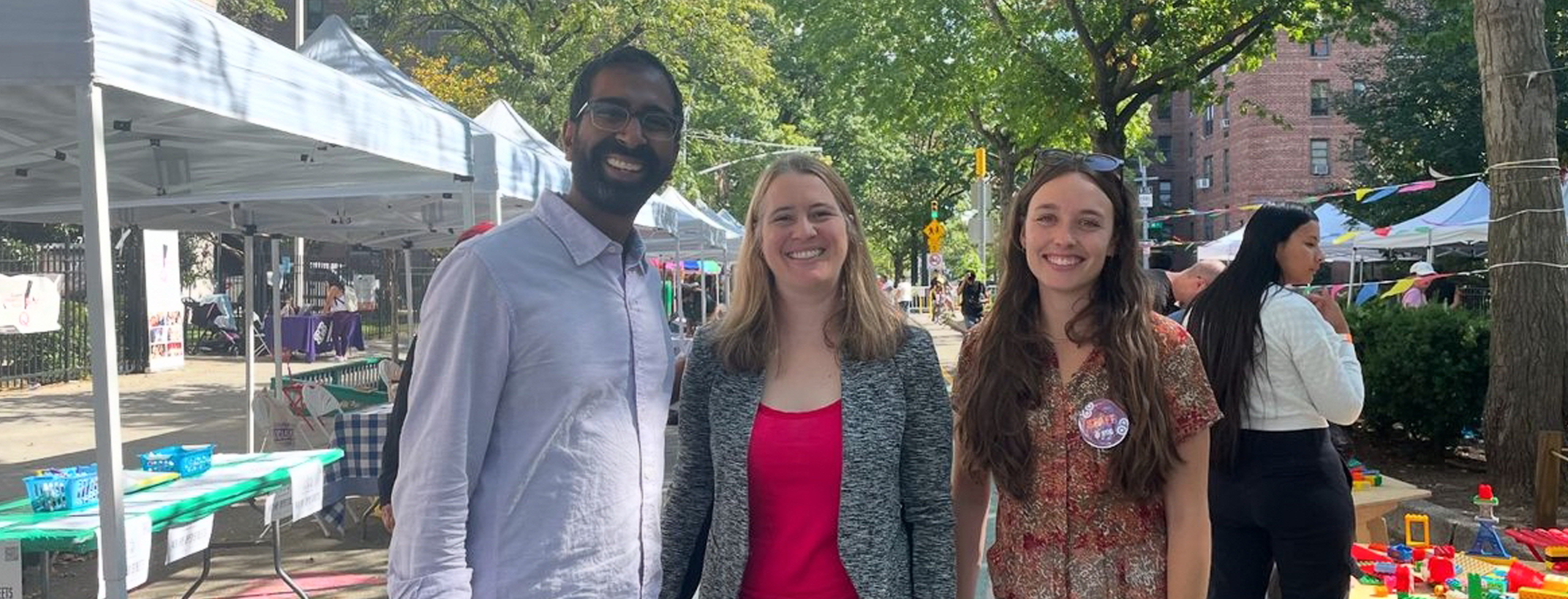 Three individuals smile at the camera with white tents behind them at an NYC neighborhood street fair