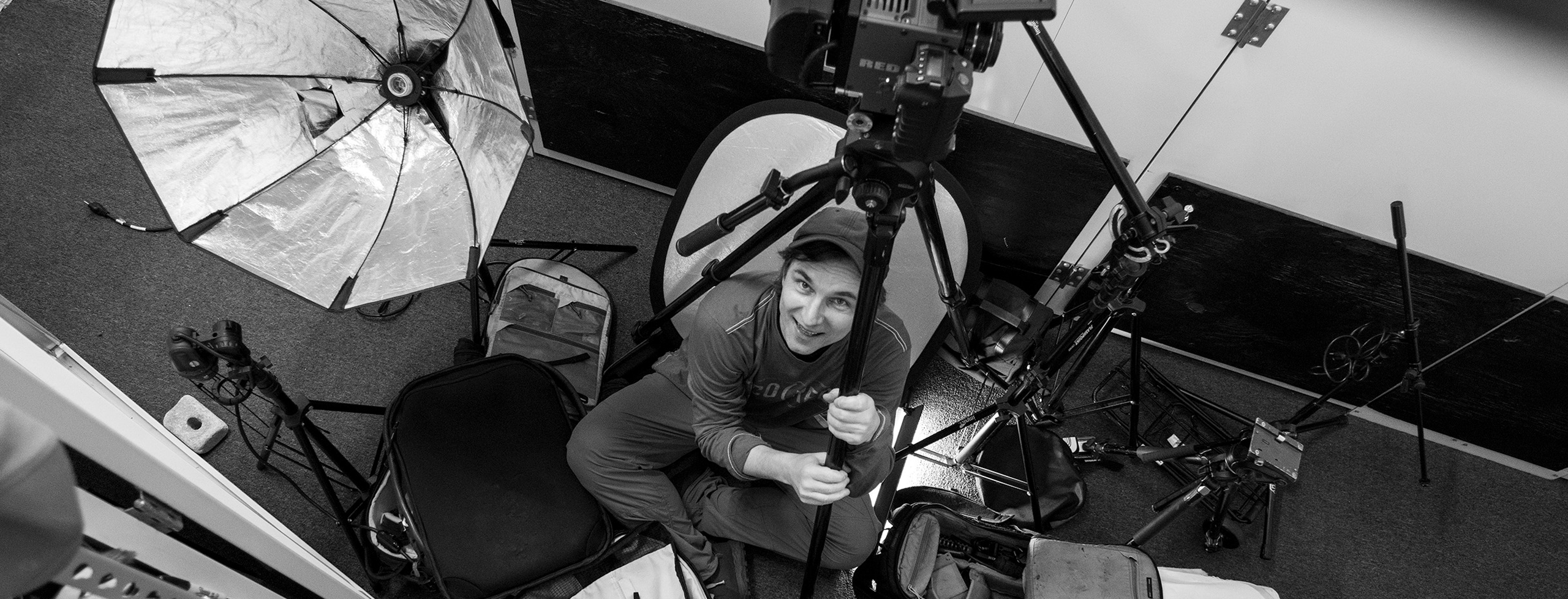 Black and white photo of a person sitting among various camera and video equipment, viewed from above.