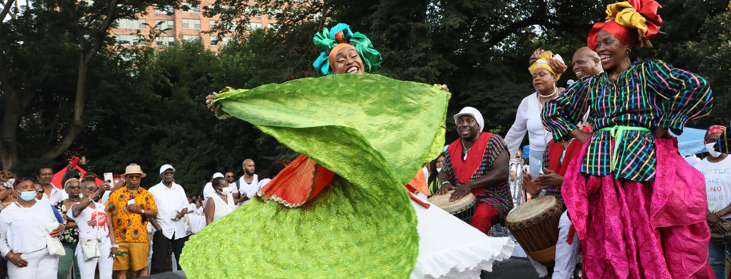 A solo dancer, center stage in a bright green dress, white petticoat and green and gold headtie. She is turning with her arms and skirt upraised. She is surrounded by female dancers in hot pink costumes.