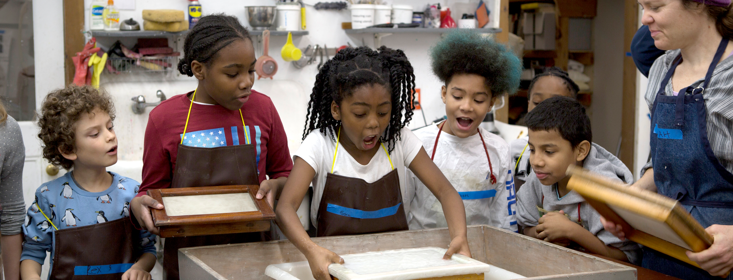 Five fourth-grade students smile with delight as one pulls a sheet of handmade paper from a vat of water. Their instructor, Hannah O'Hare Bennet, stands to the side supervising.