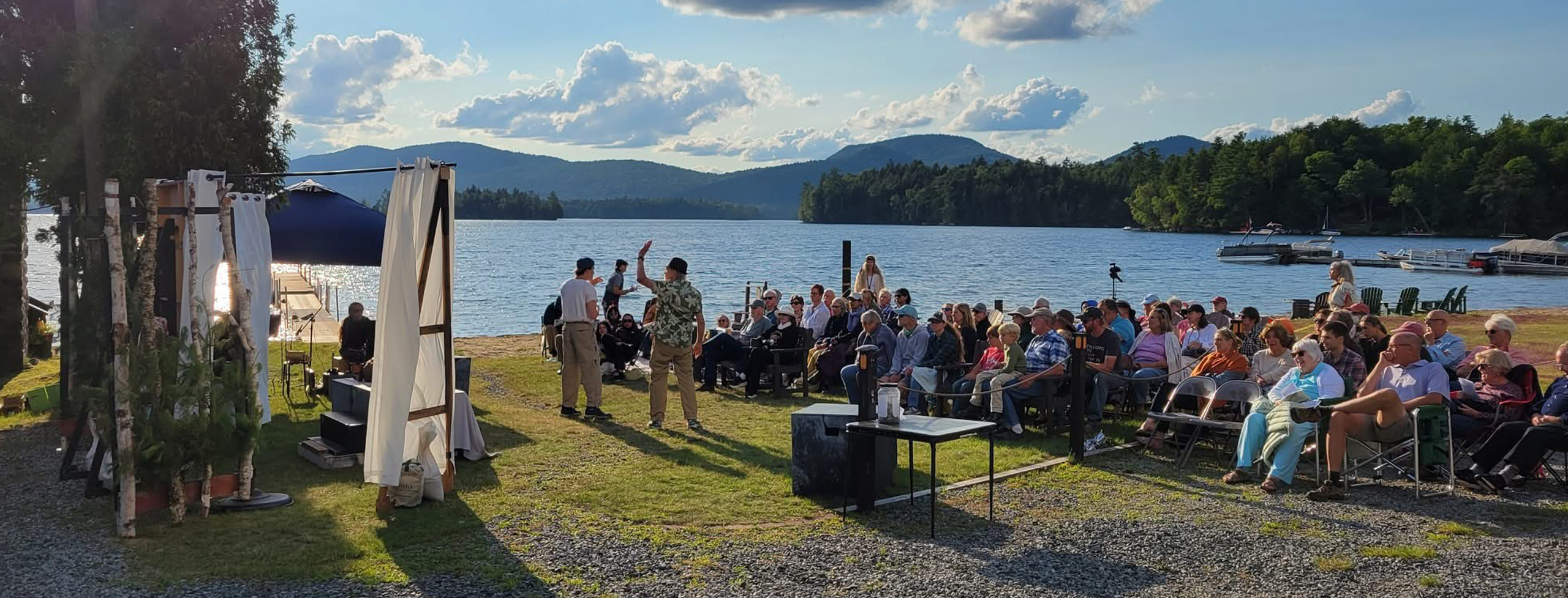 This is a picture from the 2025 Shakespeare performance of "As You Like It" outside on ALCA's property by Blue Mountain Lake. You can see the mountains and lake, with the blue sky and clouds in the background, with the audience and production in the front of the picture.