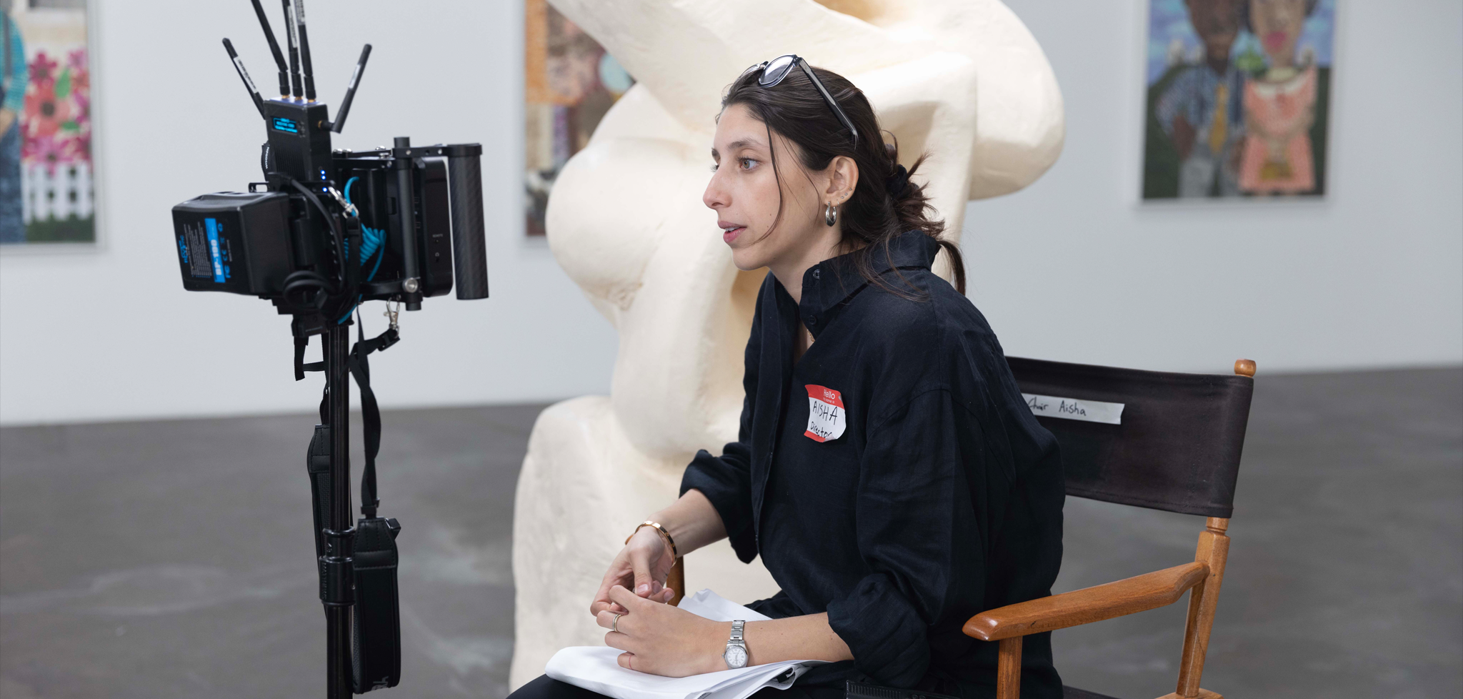 Aisha Amin seated in a director's chair in an art gallery setting, looking through film equipment