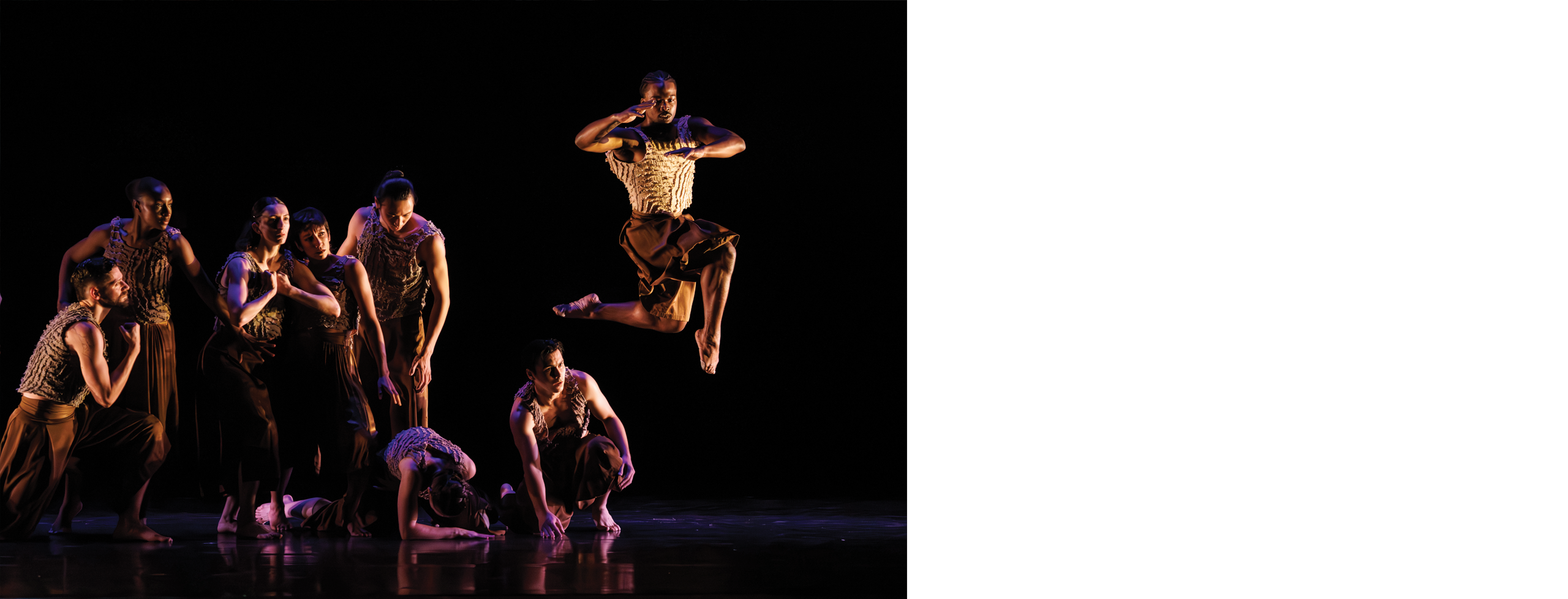 Nine dancers performing on a stage, one captured in air as the others are in different stages of movement.