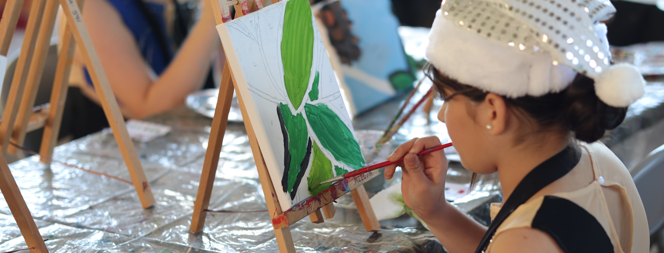 A young girl with a white, Santa-like hat on holds a paint brush in hand, working on a small canvas depicting bright green leaves