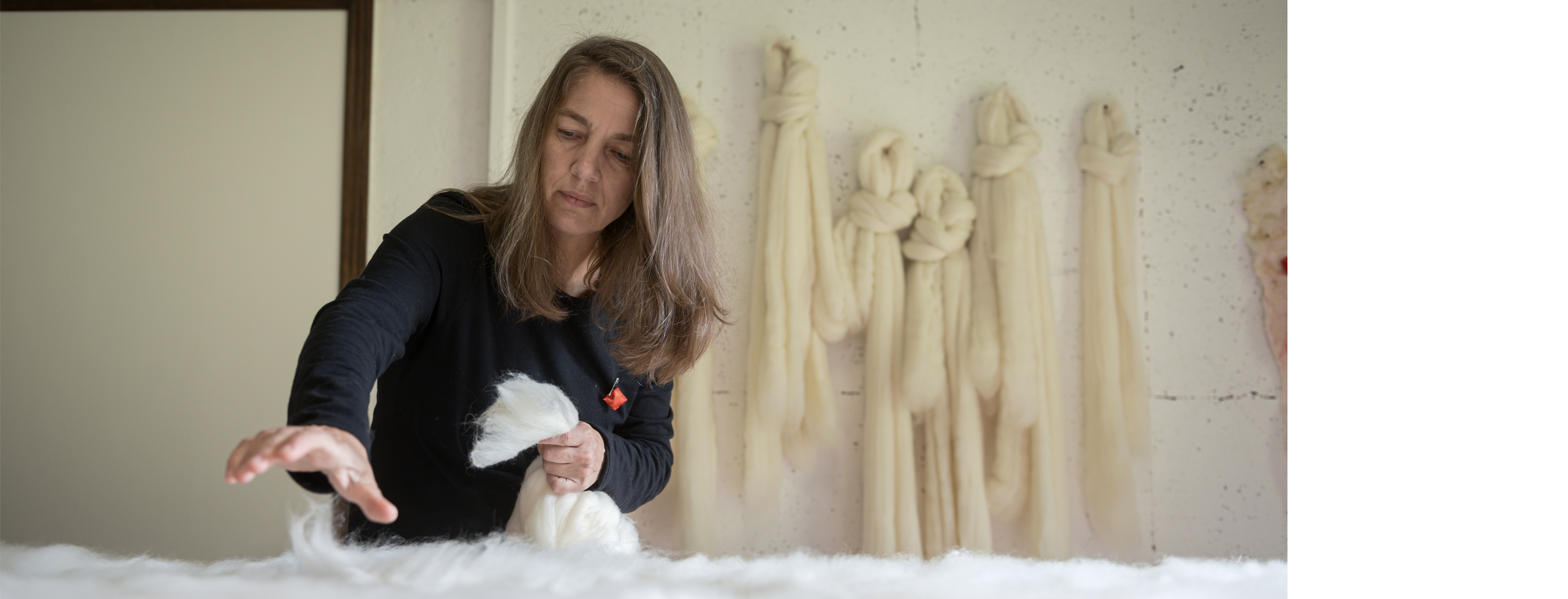 In a studio, a woman carefully layers soft white wool on a table, with long bundles of pale fiber hanging neatly on the wall behind her.