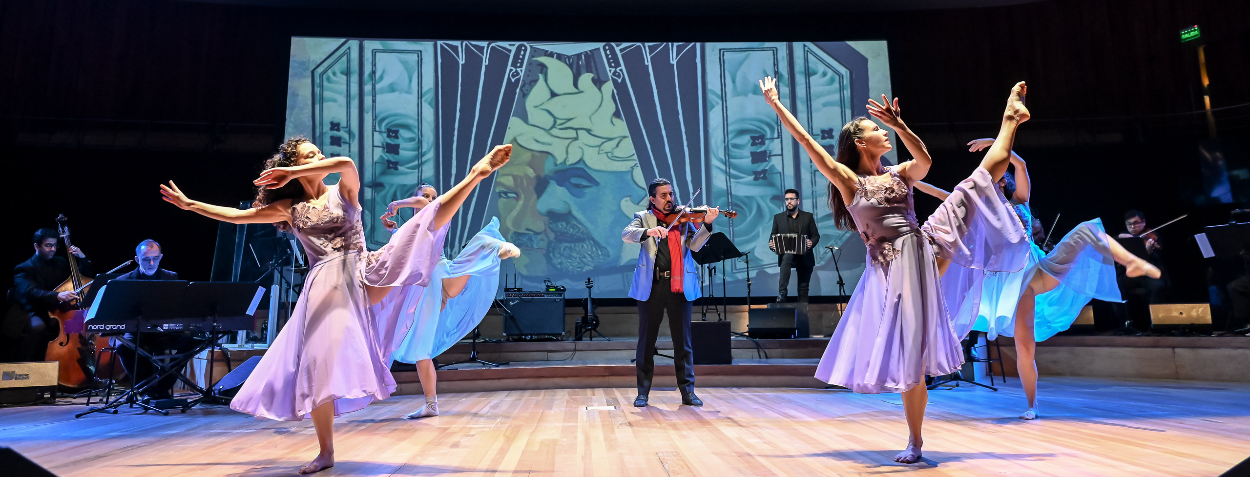 Dancers and musicians on a stage, dancers with one leg extended into the air at an angle, the opposite arm raised in the air