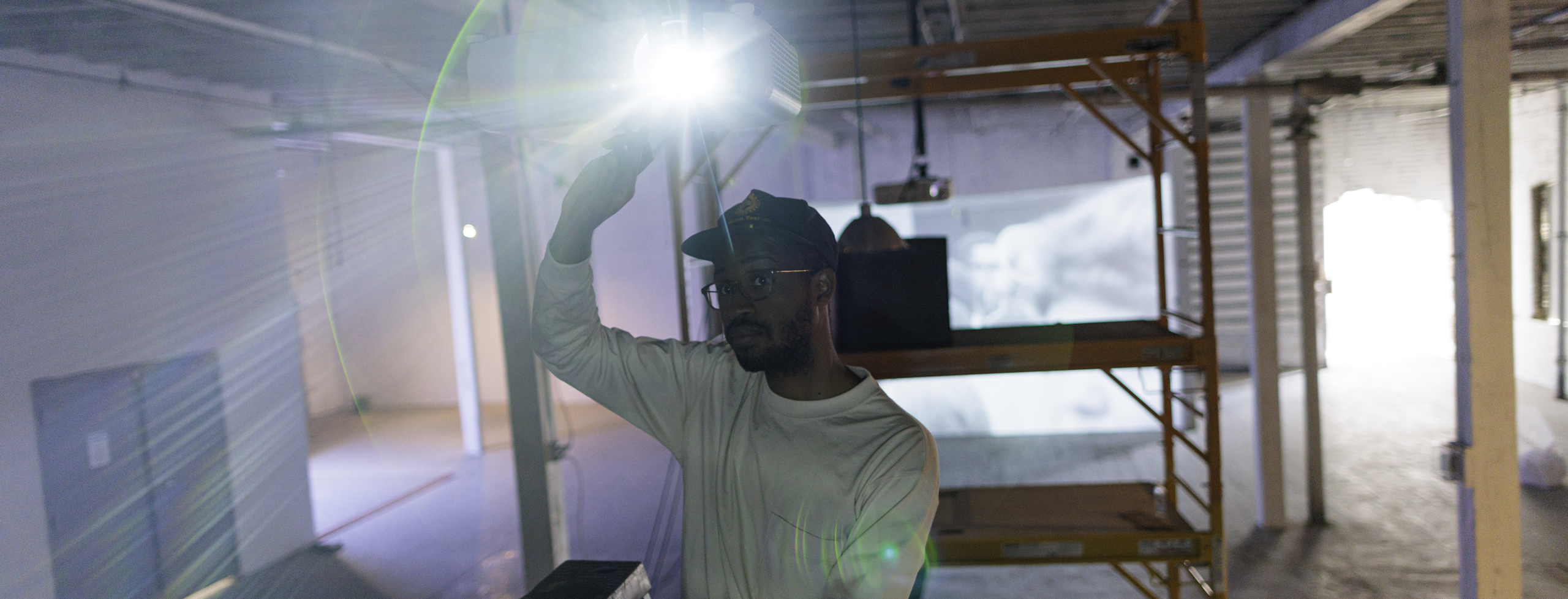 A person wearing a hat and glasses stands on a step ladder in an industrial interior, adjusting a projector that shines brightly toward the camera. Scaffolding and exposed beams are visible in the dimly lit space.