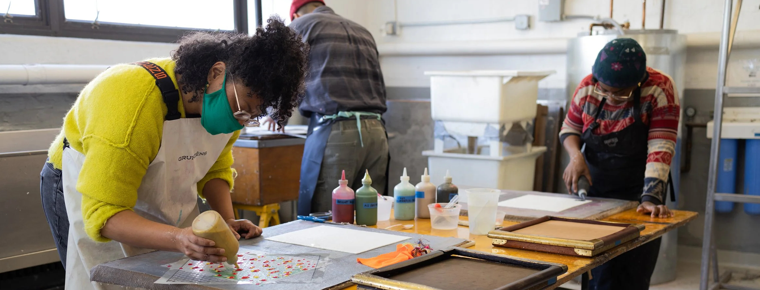 Three artists working in a studio at various stages of papermaking. Campbell wears a chartreuse sweater and leaning over a table while standing, painting with a pigmented paper pulp filled bottle.