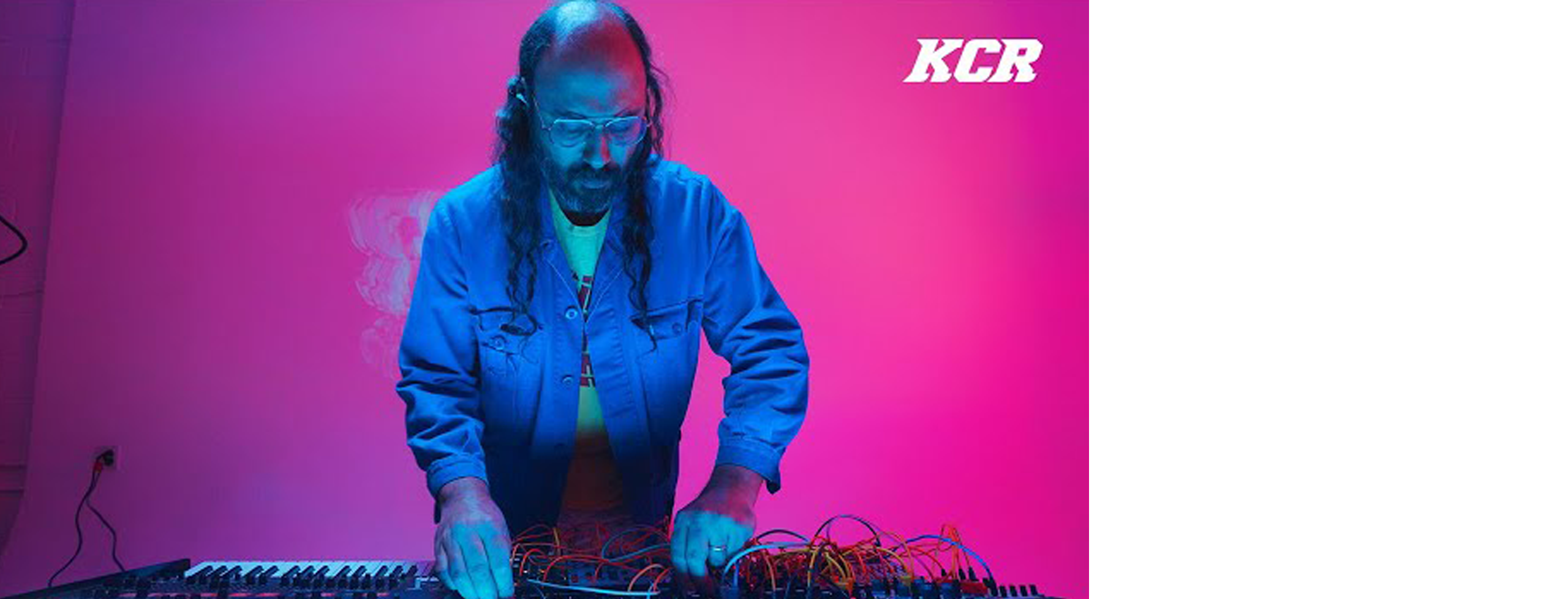 A South Asian man with a beard and long hair, performing a modular synthesizer in front of a purple backlit wall