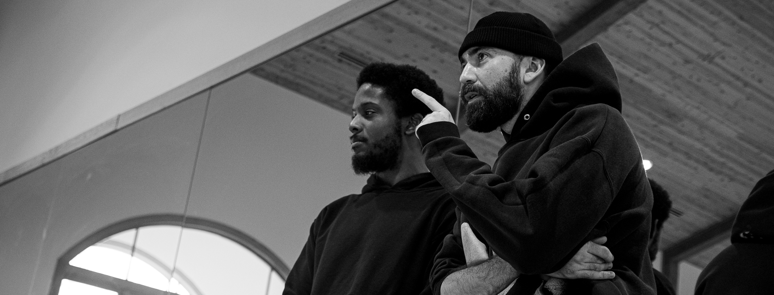 A white man and a black man stand with their back to a mirror conversing on how to move forward with rehearsal. The white man points towards the center of the room.