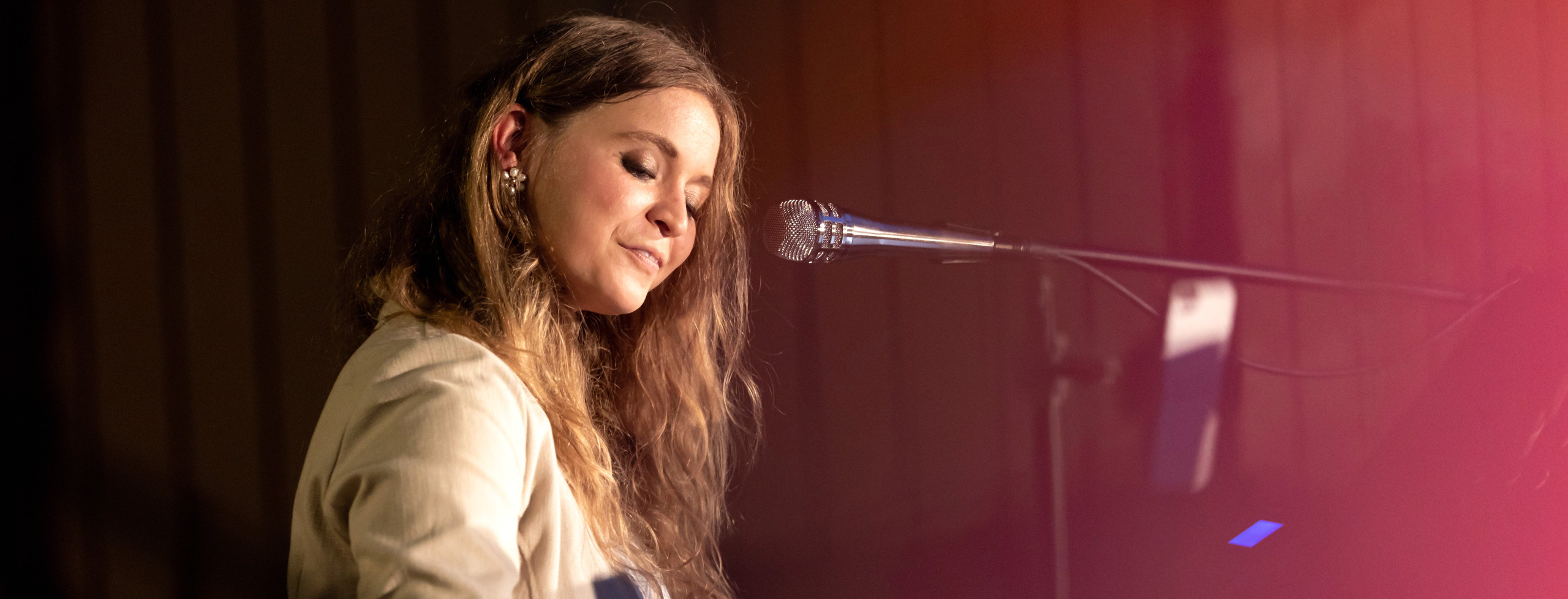 A Latvian girl with bright hair and sand color clothes playing piano and singing.