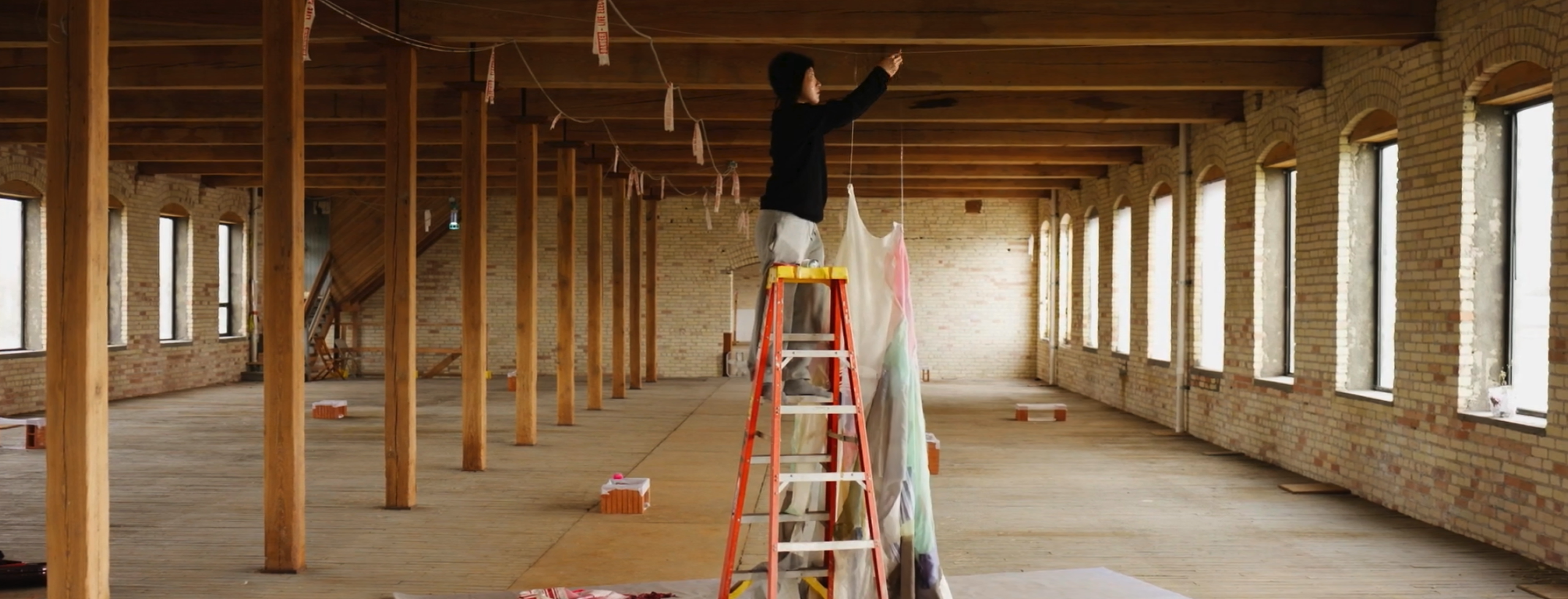 An artist stands on a ladder inside a large wooden interior space while installing hanging textile elements. Fabric pieces and materials are spread across the floor as part of the installation process.