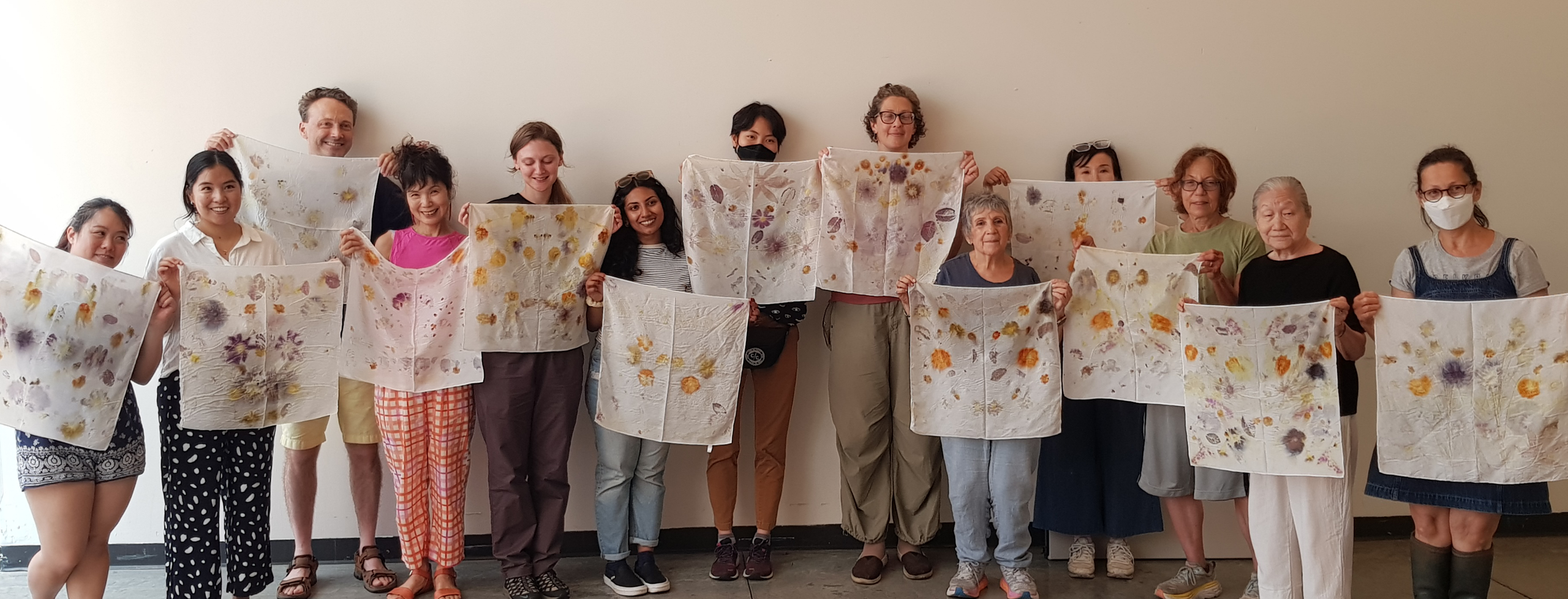 An Indoor group photo showing thirteen people, each holding up a square shaped fabric. Standing side by side, everyone is standing against the white wall, facing the camera while each showing the printed textile artwork he/she/they made.