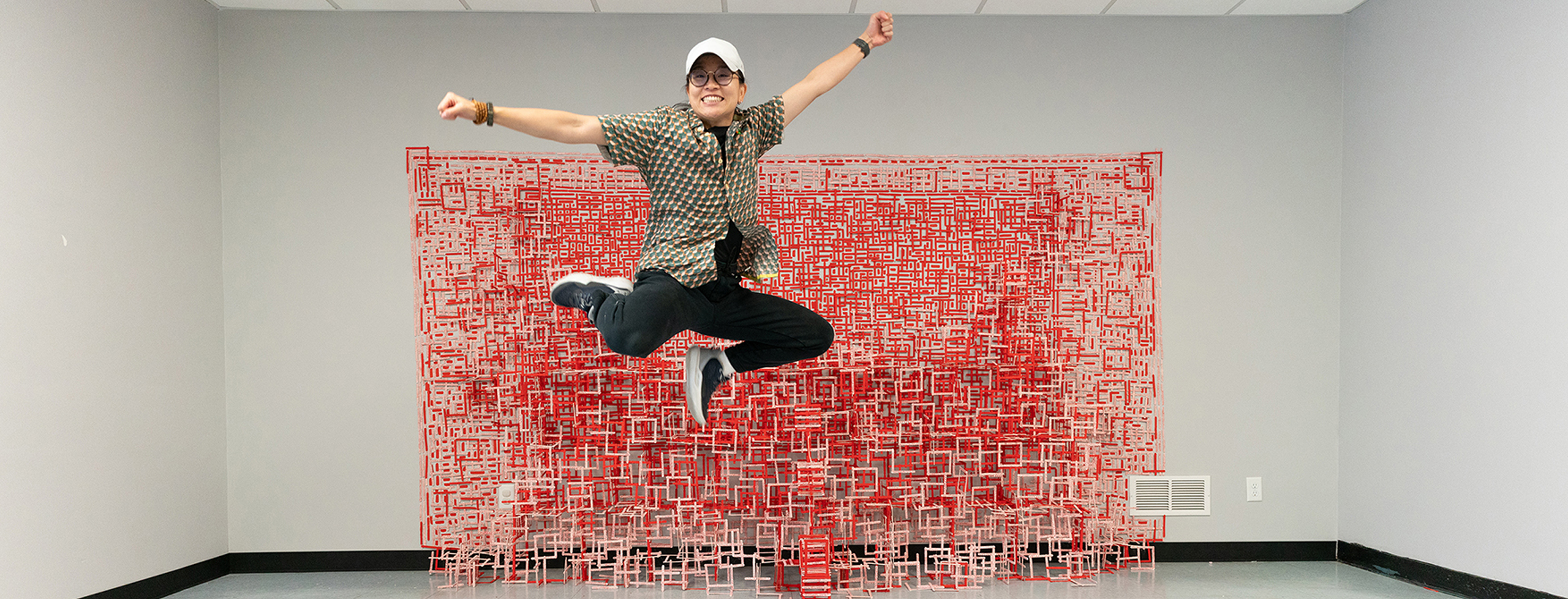 Artist Ellen Xu jumps in front of a large red installation made from hundreds of painted popsicle sticks forming a layered architectural structure.