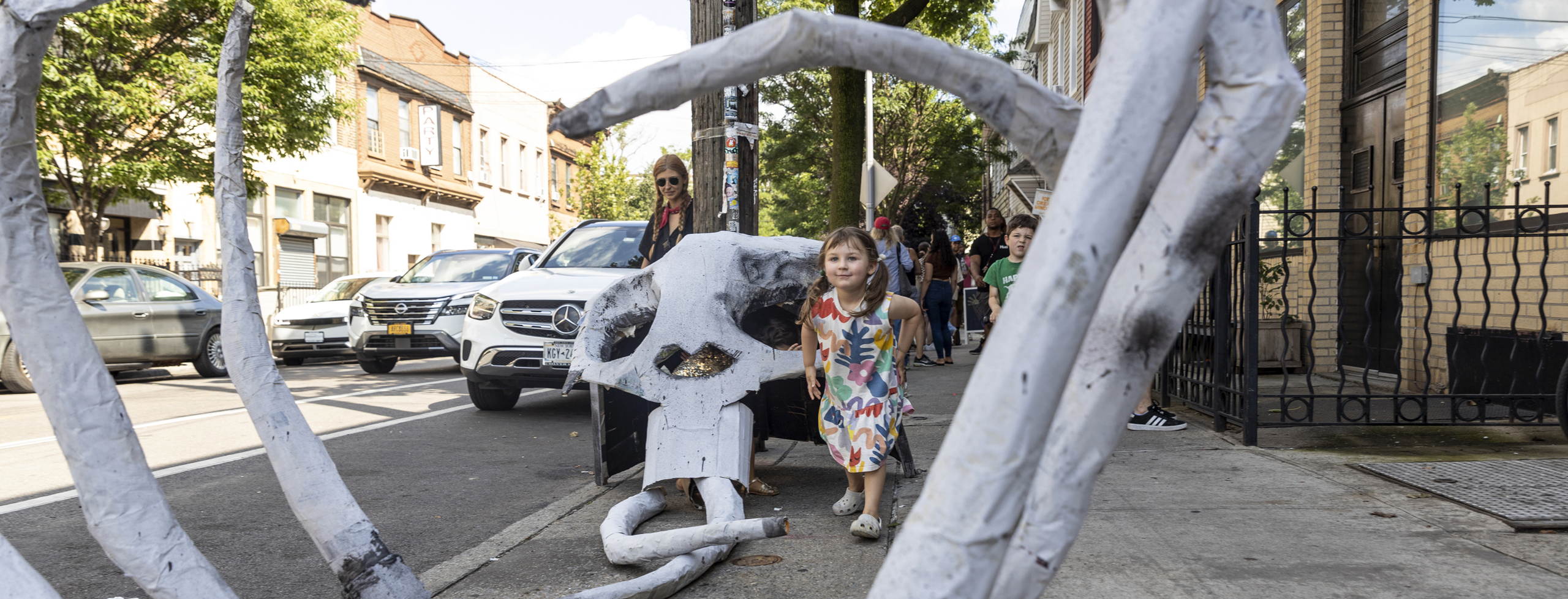 A large white sculpture shaped like a skeletal rib cage rises from a small tree bed along a city sidewalk. The curved ribs arch outward, forming a tunnel-like structure that frames the view toward the street. A young child walks past the sculpture while other pedestrians and parked cars line the block behind them.