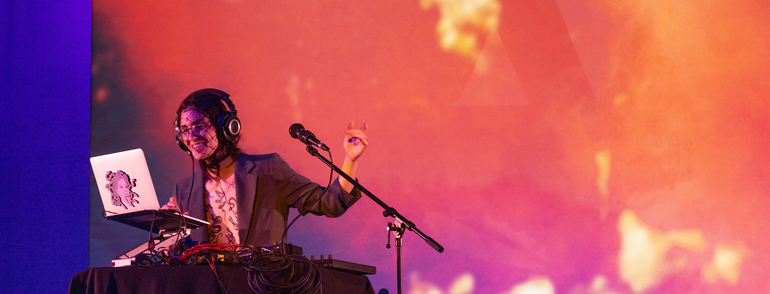 A musician wearing a fishnet face covering performs electronic music in front of a large projection of fire.