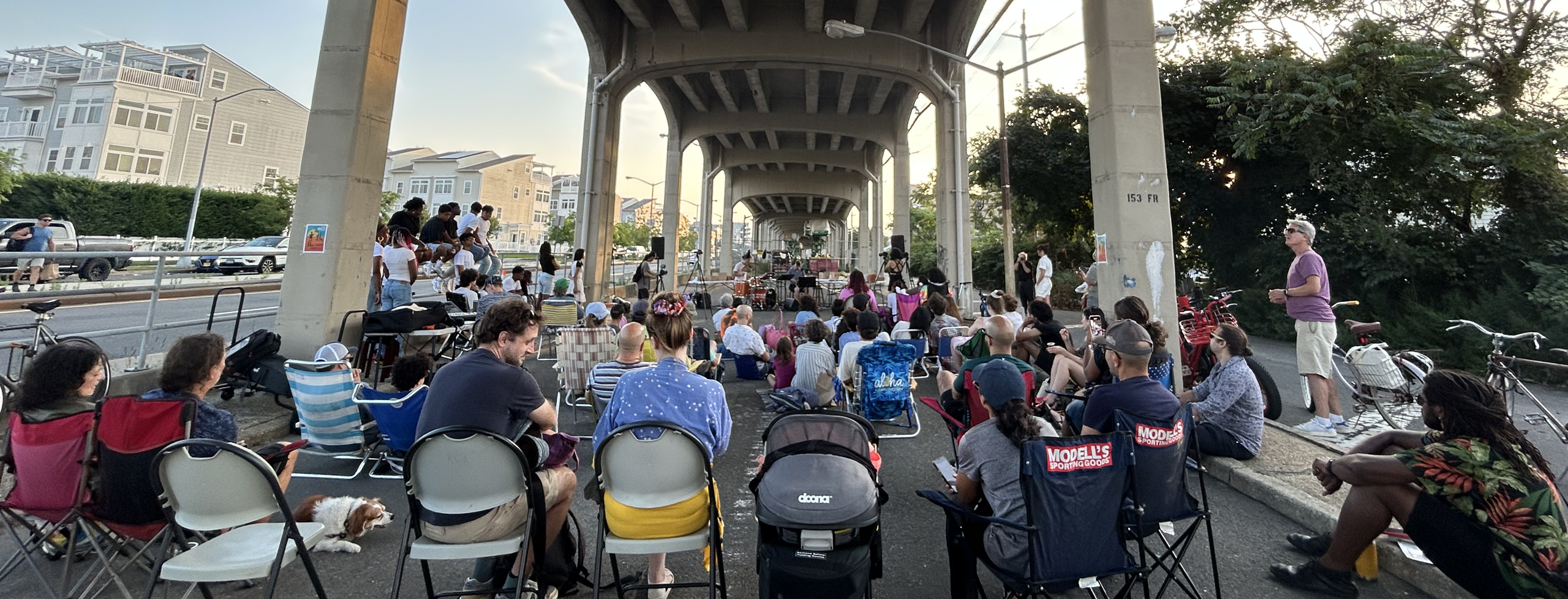 There is a crowd of people in lawn chairs under the elevated A train tracks. Percussionists are performing in the front and dusk is just about to begin.
