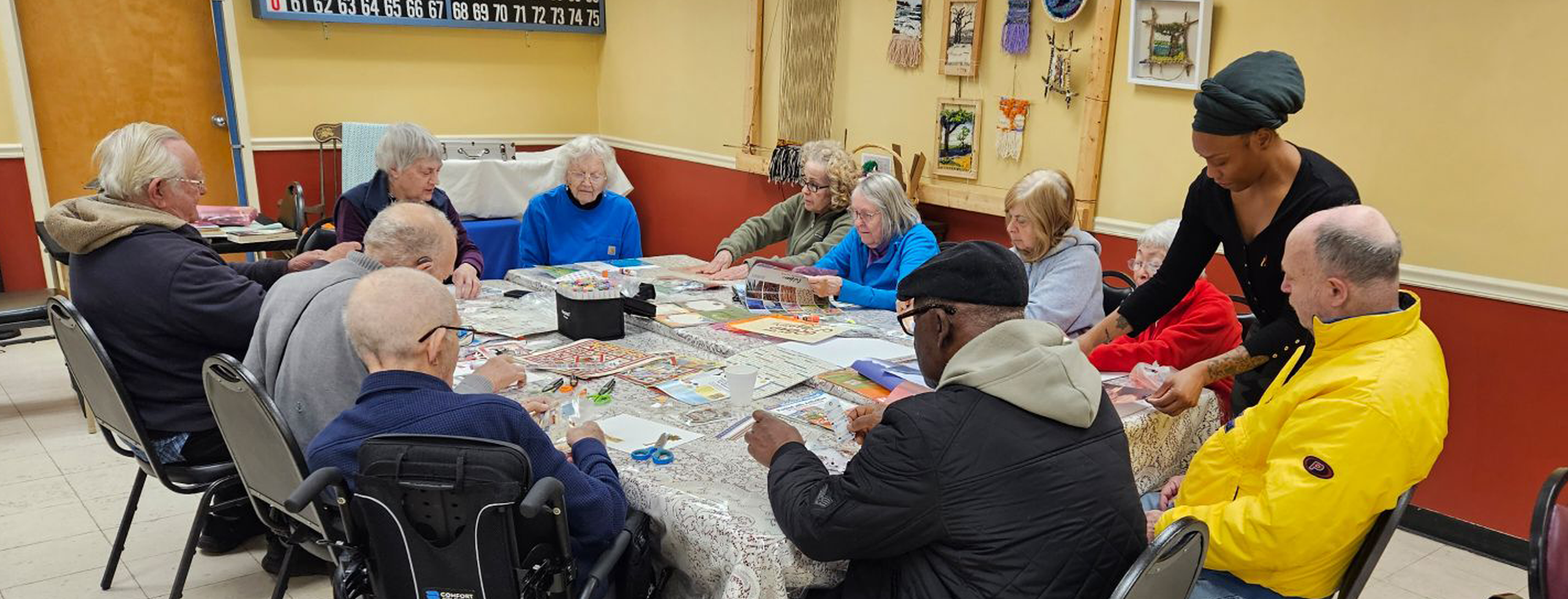 An individual stands, helping someone at a table of senior citizens who are making collage-based artworks