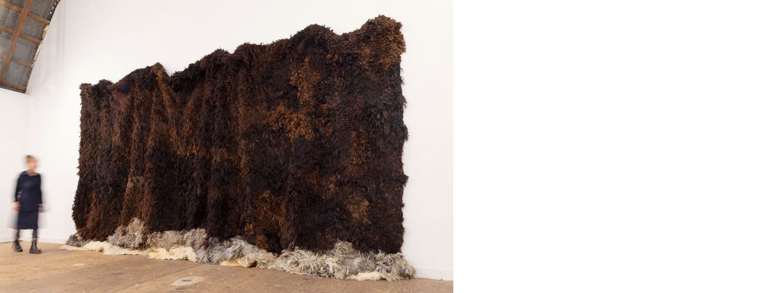 Photo of a blurred person walked by a large brown woolen work hanging from the white wall of a gallery--for scale and context.