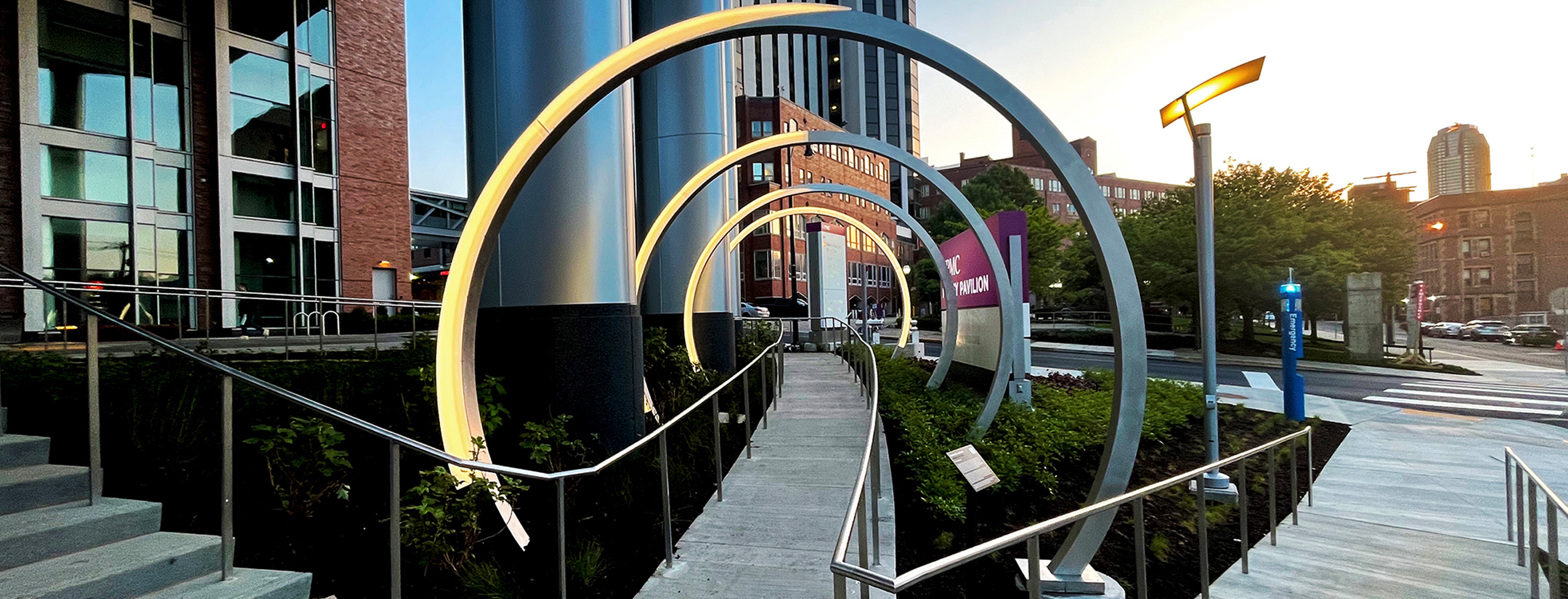 An outdoor installation of four 10-foot stainless steel, acrylic, and LED light archs positioned along a straight pedestrian pathway in a medical plaza in Pittsburgh, PA. Description: The Lunar Portal is a 10-foot stainless steel and acrylic archway installation, with each arch representing a phase of the Moon along a pedestrian pathway. Its custom LED lighting system adjusts in sync with the lunar cycle, encouraging viewers to reflect on how moonlight influences daily life and well-being.