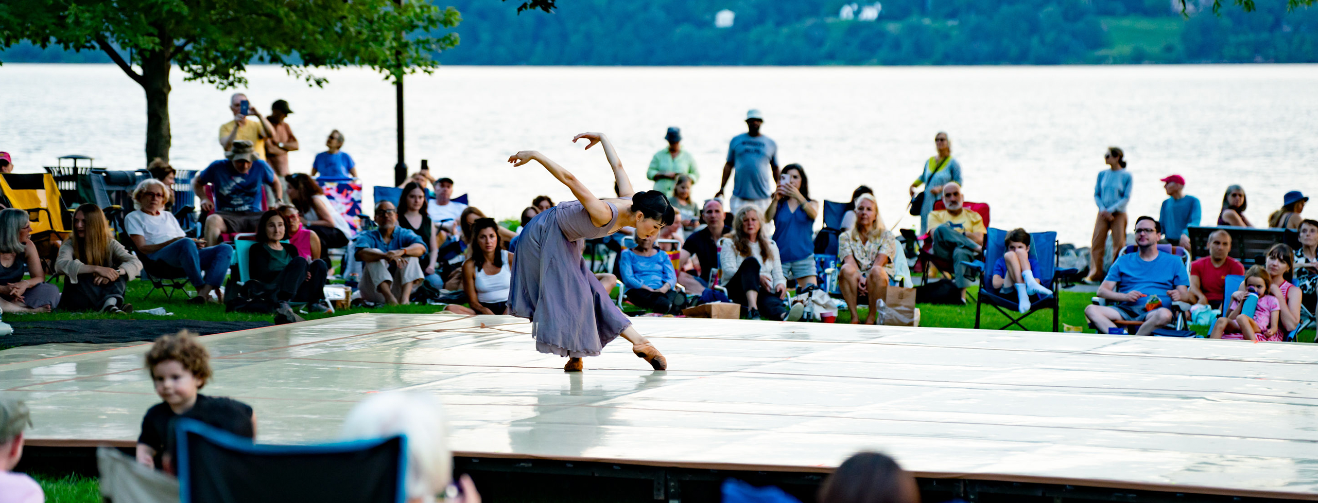 A single dancer appears on a stage set against the backdrop of the Hudson River (Dobbs Ferry Waterfront Park), people set up in lawn chairs surrounding the stage.