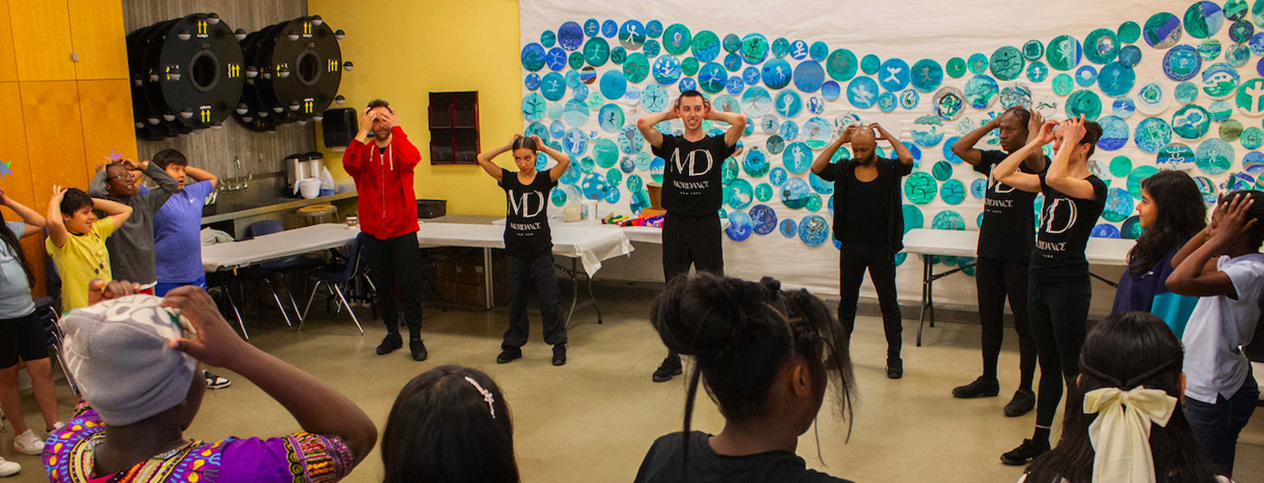A dance lesson takes place in a classroom-like setting, everyone with their arms raised and hands on their head
