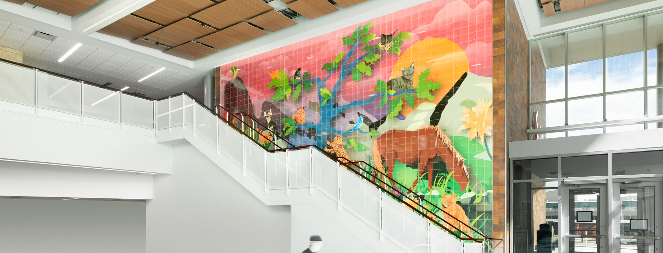 A colorful ceramic tile mural in a building lobby depicts animals—including cats, birds, and a horse, set in a vibrant landscape of trees, flowers, and a large sun, creating a joyful and welcoming atmosphere.