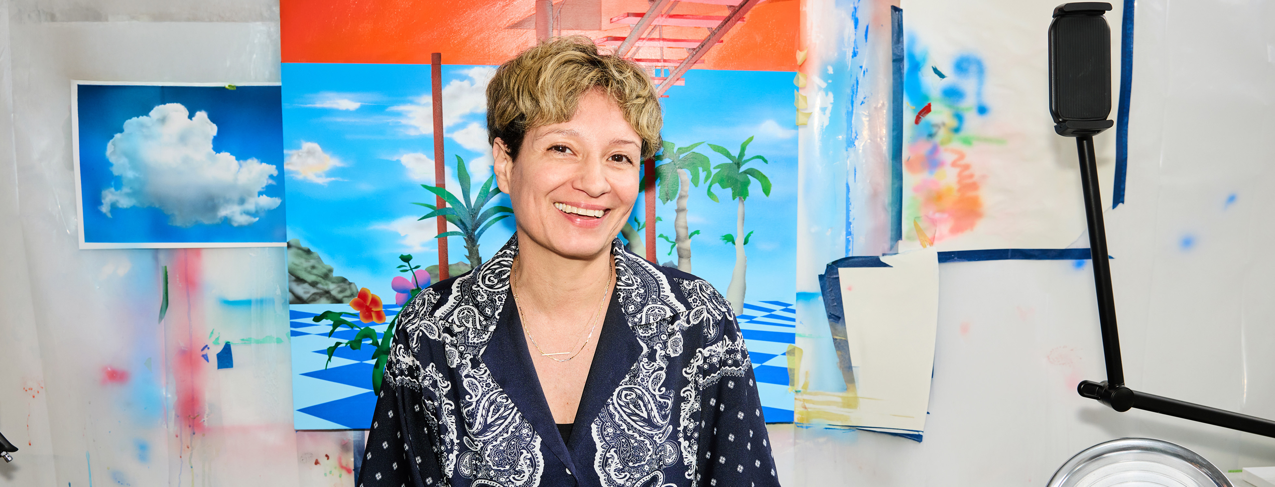 Priscila De Carvalho sits in her studio in front of a colorful painting in progress, featuring palm trees, a geometric floor, and architectural elements against a vivid sky.