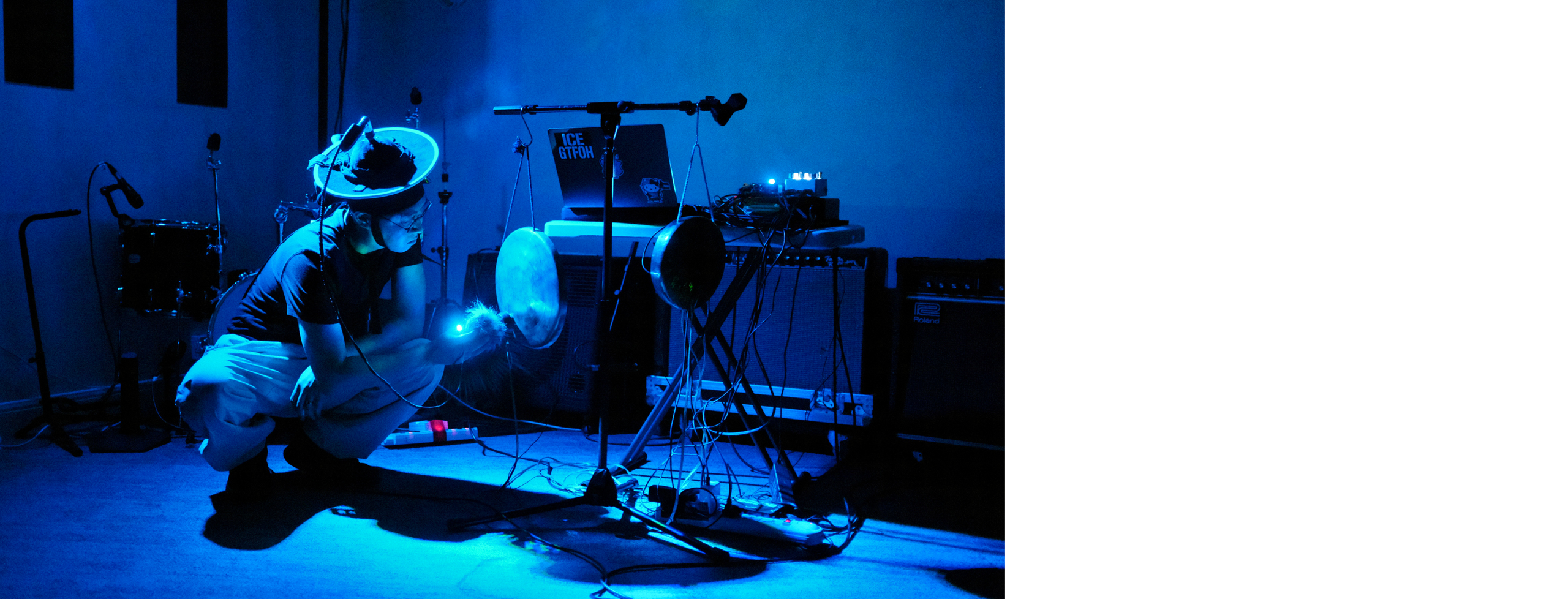 One performer is squatting and holding a microphone strung to a hat up to a gong that is hanging from a stand in blue ambient lighting.