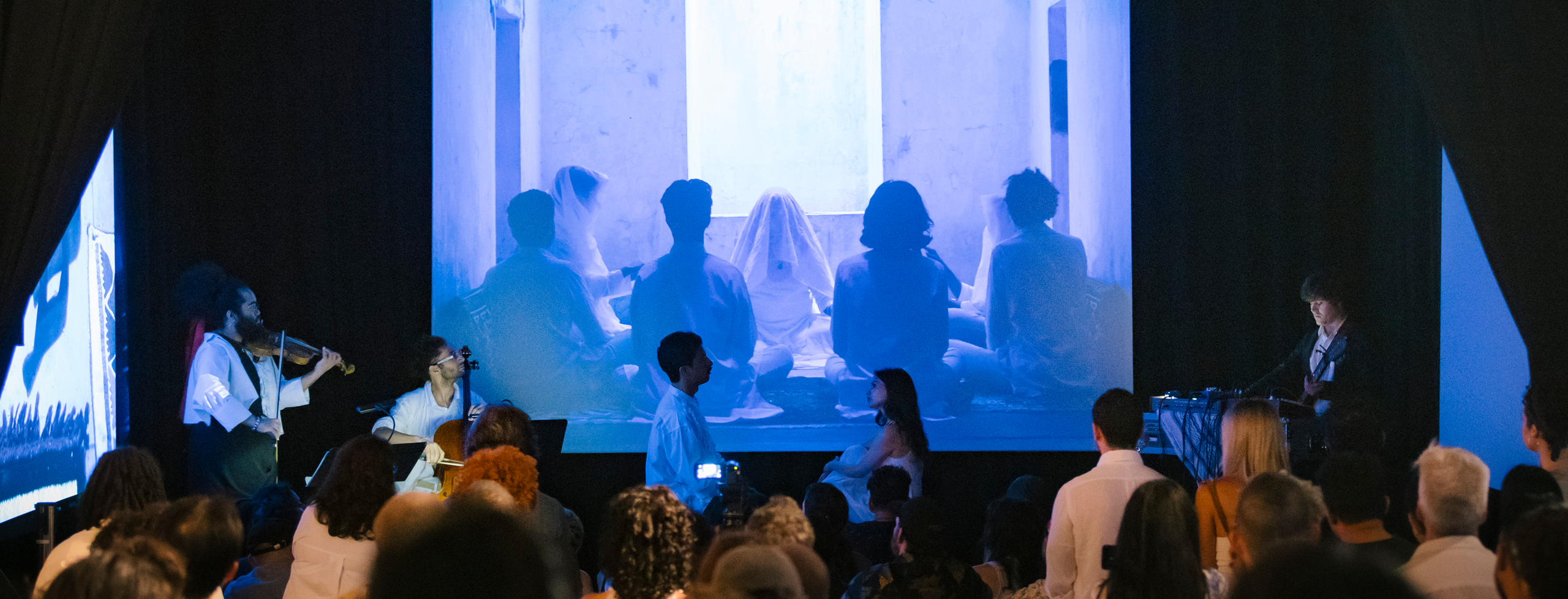 An audience sits facing performers (left to right: Darian Donovan Thomas on violin, Zeelie Brown on cello, Zain Alam on vocals, Zeerak Ahmed on vocals, and Warren Hildebrand on guitar/electronics) in front of a large three-channel video installation in a dark gallery. The central projection shows a group of silhouetted figures gathered around a table, while matching projections extend onto the side walls.