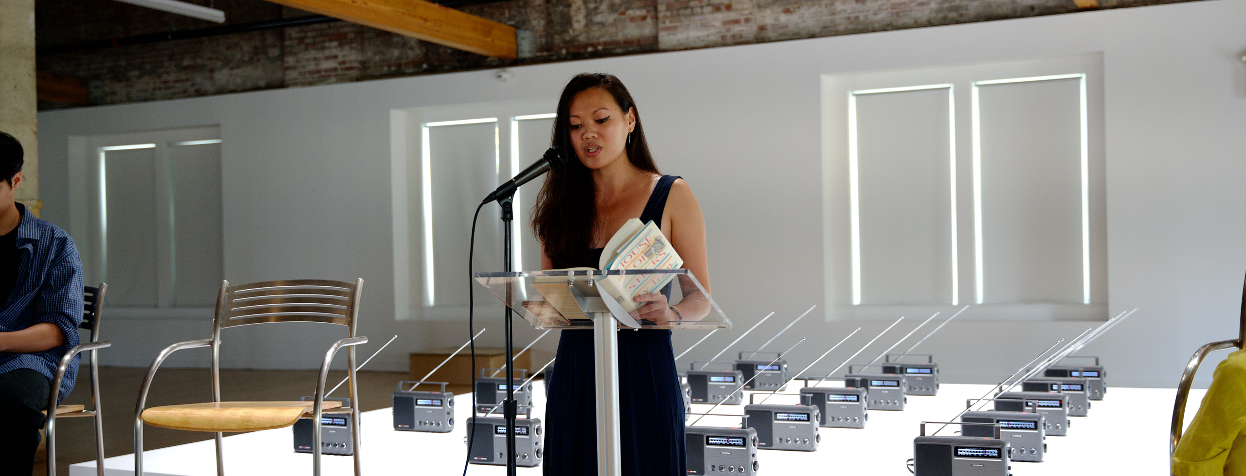 Author Ly Tran in a navy blue dress reads from her memoir House of Sticks at a clear podium with a microphone, holding an open copy of her book. Behind her is a large art installation of vintage radios arranged on white tables in a spacious gallery with exposed brick walls and tall windows.