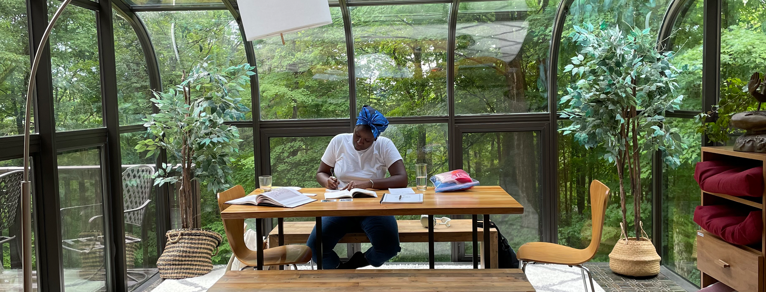 Alt Text: Angel Nafis sitting at a table at work on poetry in a glass atrium, the woods of Connecticut in the background