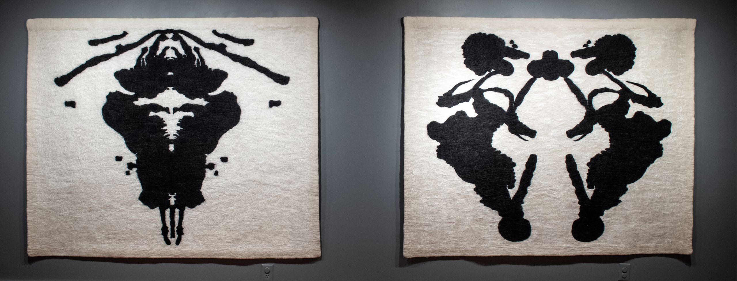 The dark gray wall holds two large white textiles with symmetrical black patterns. Track lighting casts individual spots of light onto each textile.